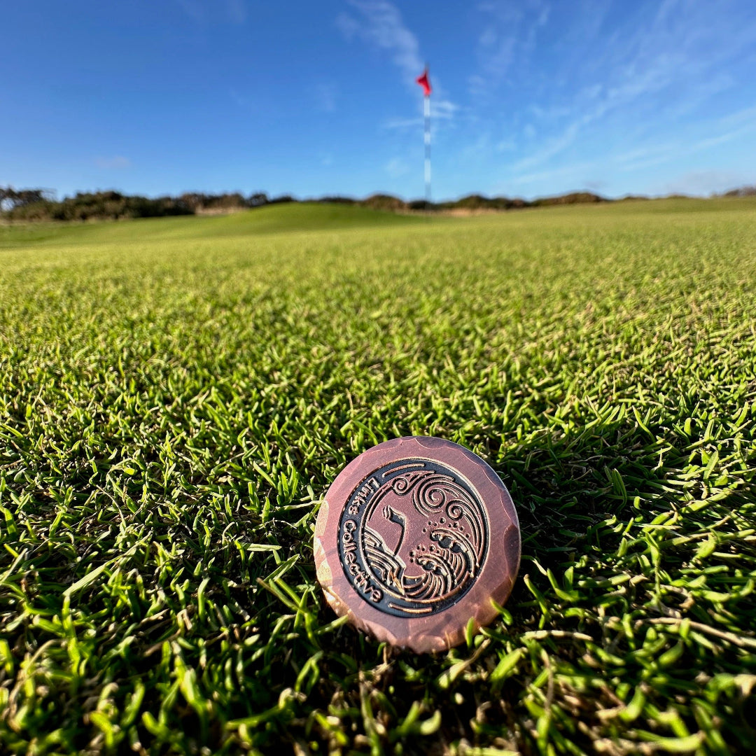 An iron golf ball marker with a custom logo engraved on it, placed on a green on a golf course with a blurred background.