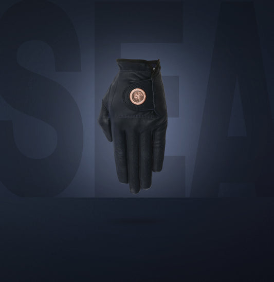 A blue cabretta leather golf glove with a forged metal ball marker attached, set against a dark background with the collection name 'SEA' visible in the background.